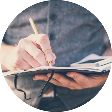 Nahaufnahme einer Person, die mit einem Bleistift Notizen in ein aufgeschlagenes Notizbuch schreibt, im Hintergrund unscharf ein Smartphone.