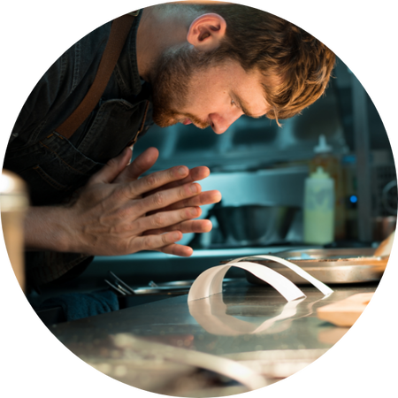 Ein konzentrierter Koch in Schürze beugt sich über einen Arbeitstisch in der Küche, die Hände gefaltet vor sich, neben zwei Bestellzetteln.