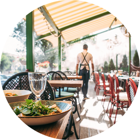 Sonnige Restaurantterrasse mit gedeckten Tischen im Vordergrund, ein Glas und ein Salatteller auf dem Tisch; im Hintergrund geht ein Kellner in Schürze durch den Aussenbereich.